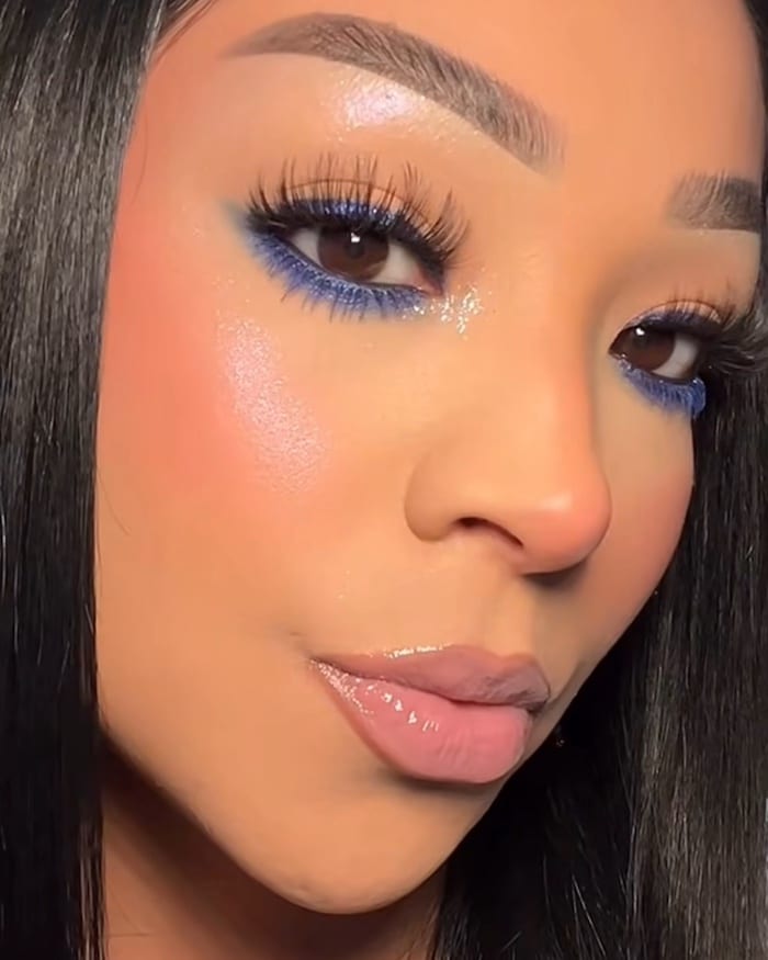 A person with straight black hair wears glossy nude lipstick, glowing peach blush, shimmery eyeshadow, bold eyebrows, and dramatic false lashes with vivid blue eyeliner on the lower lash line for a vibrant style.