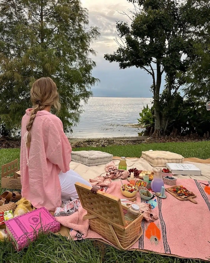 A woman in a pink shawl sits on a colorful blanket at a lakeside picnic, reminiscent of a Girls Life June/July issue cover, surrounded by food, cushions, plush toys, and nature’s calm under a cloudy sky.