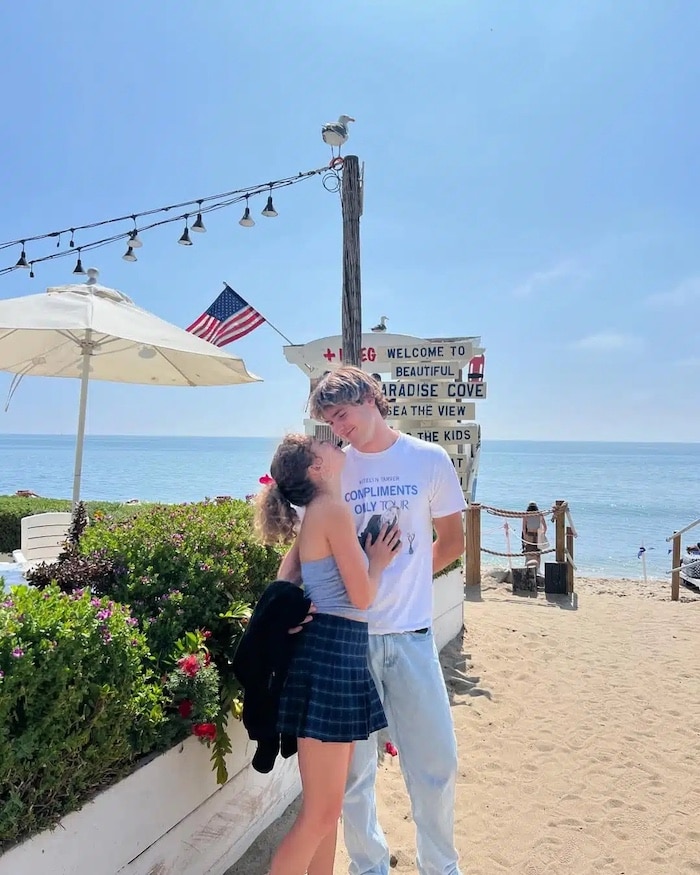 A couple stands close together on a sunny beach near a lifeguard station; the woman is smiling up at the man. The scene, reminiscent of a Girls Life June/July Issue cover, features an American flag, string lights, umbrellas, and the ocean.
