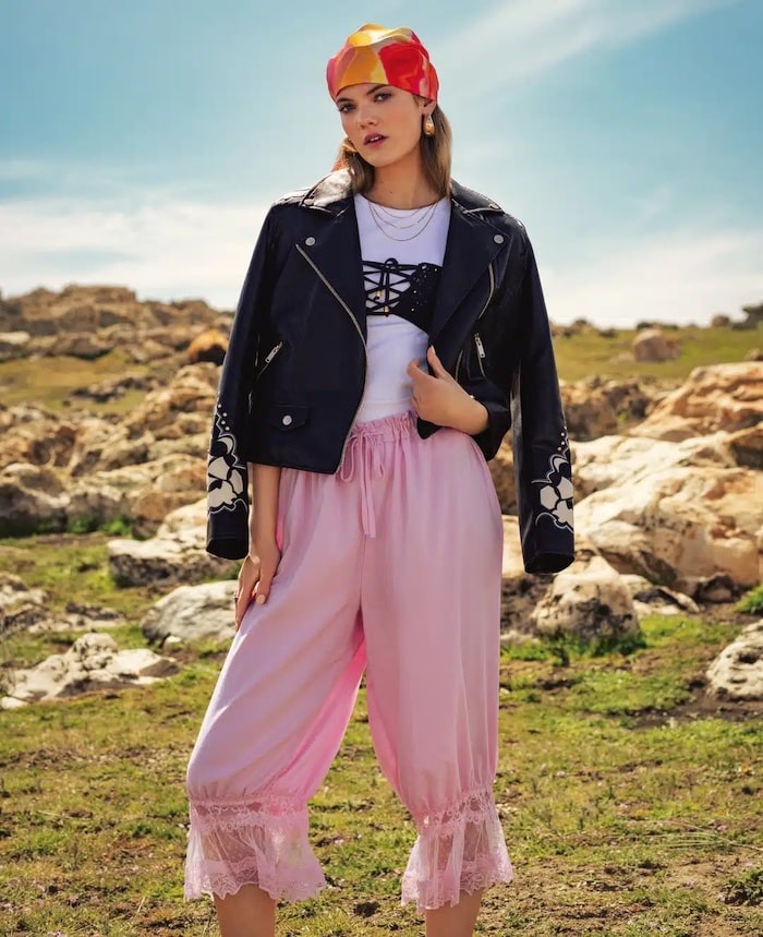 A woman stands outdoors on grassy terrain with rocks, wearing a red and orange headscarf, black leather jacket, white top, and pink pants with lace trim—posing confidently under a blue sky like she’s ready for the Girls Life June/July issue.