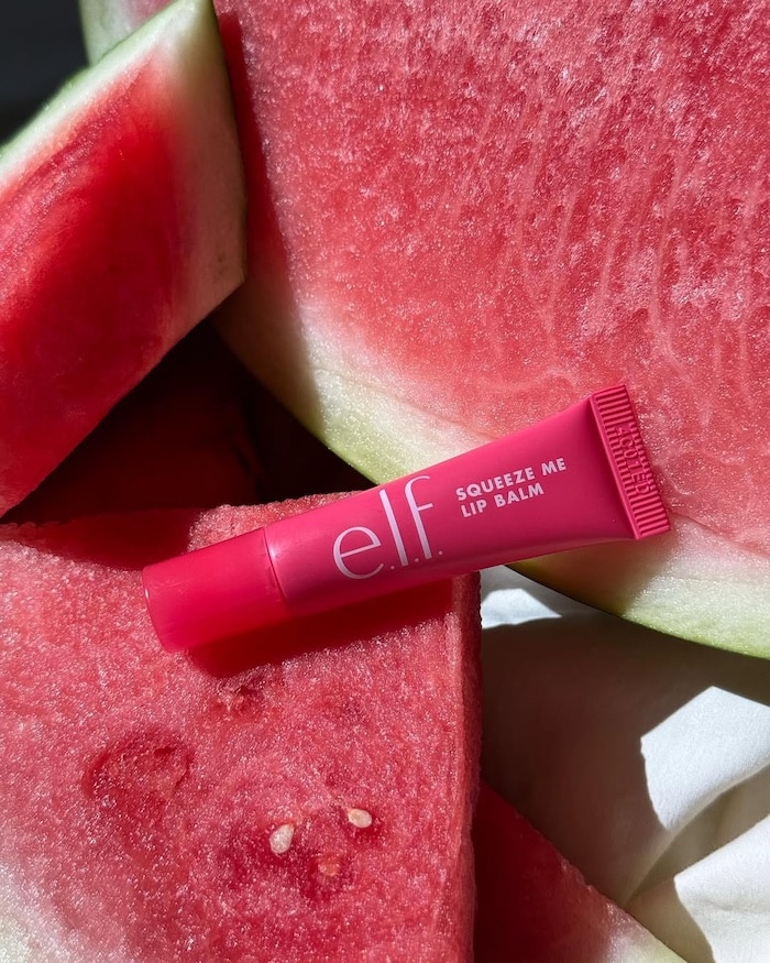 A pink tube of e.l.f. Squeeze Me Lip Balm rests on slices of fresh watermelon, the matching vibrant red packaging making it a perfect summer beauty essential.