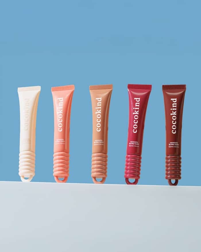 Five tubes of Cocokind lip balms—summer beauty essentials—are arranged in a row on a white surface against a blue background. Each tube features a colored cap that matches the balm shade inside.