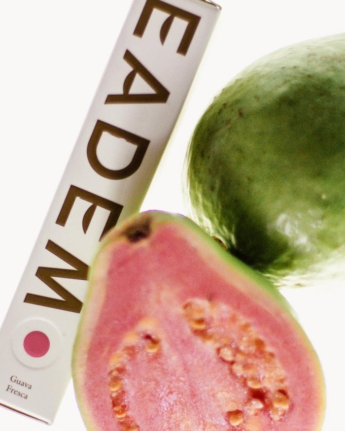 A box labeled EADEM, a summer beauty essential, lies next to a whole guava and a halved guava showing its pink flesh and seeds. The box features a circle and the words Guava Fresca—a nod to nourishing lip balms inspired by tropical fruits.