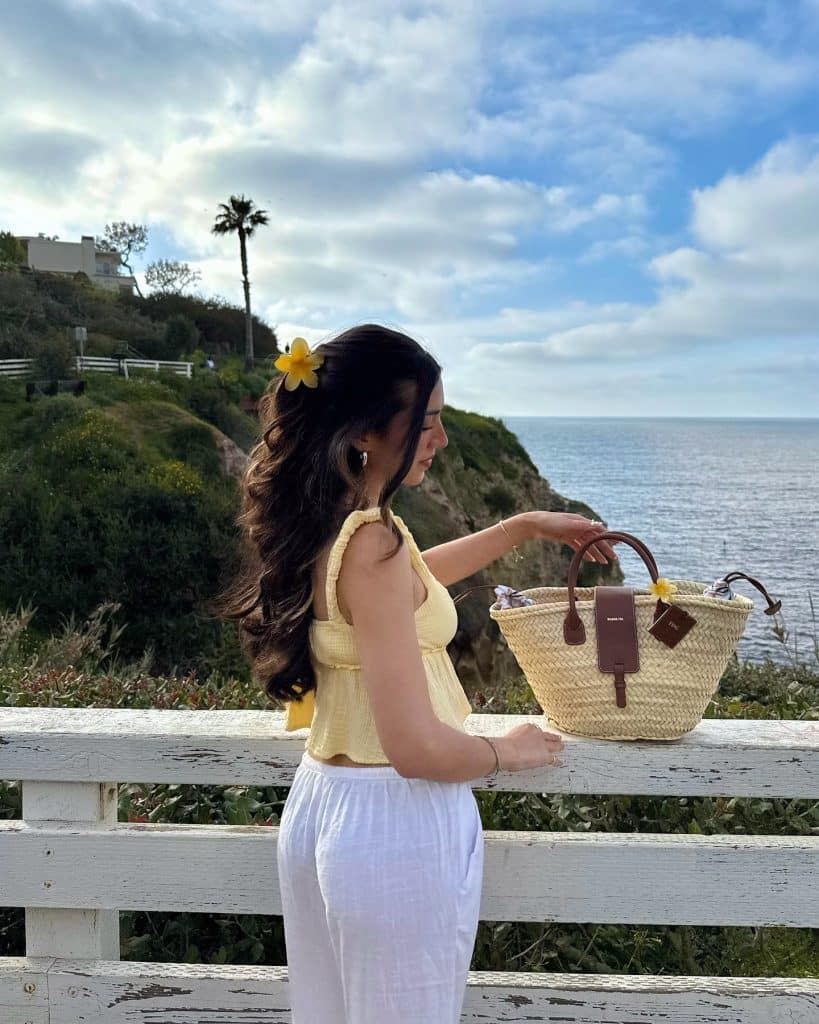 A woman with long dark hair, wearing a yellow top and white pants, stands by a white wooden fence overlooking the ocean. She holds a straw beach bag and has a yellow flower in her hair. Green hills and a cloudy sky complete this serene scene.