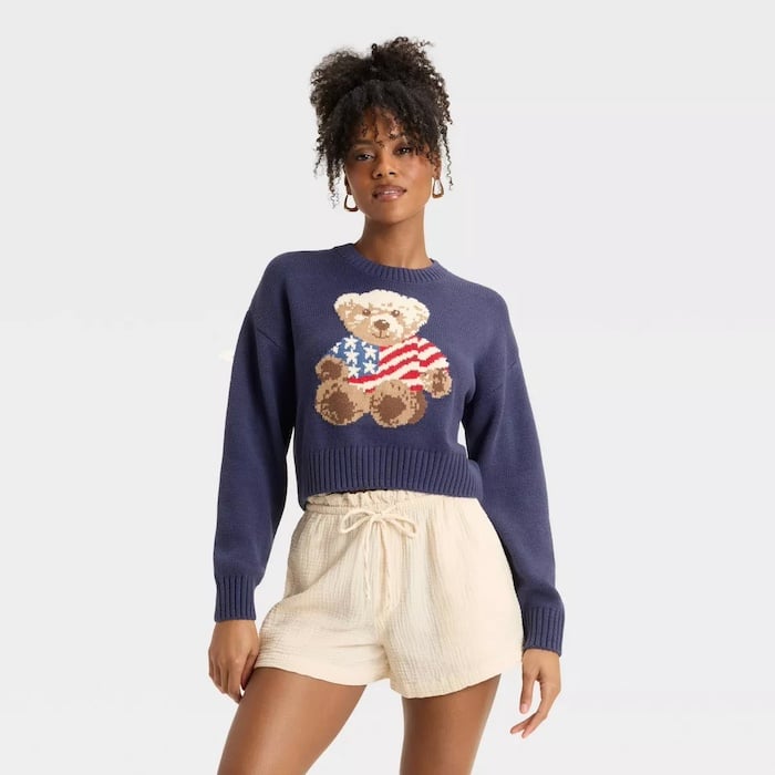 A woman wearing a navy blue sweater with a teddy bear holding an American flag—perfect for a 4th of July style guide—pairs it with cream-colored high-waisted shorts, standing against a plain light background.