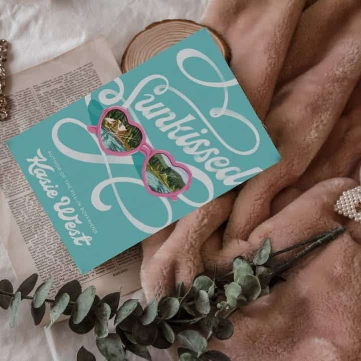 A book titled Sunkissed by Kasie West with pink sunglasses on the cover lies on a soft blush blanket, beside a newspaper, pearl hair clip, eucalyptus leaves, and a wooden coaster—perfect for fans of books like TSITP.