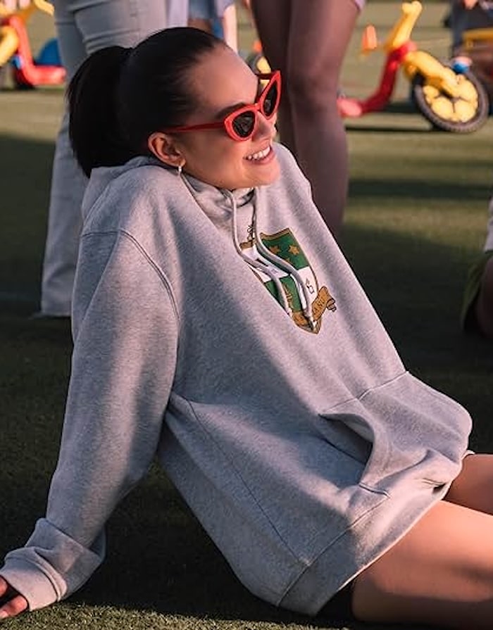 A woman wearing a gray hoodie and red cat-eye sunglasses sits on grass, smiling and leaning back with her hands on the ground—an effortless look inspired by The Summer I Turned Pretty outfits. Colorful tricycles and people fill the background.