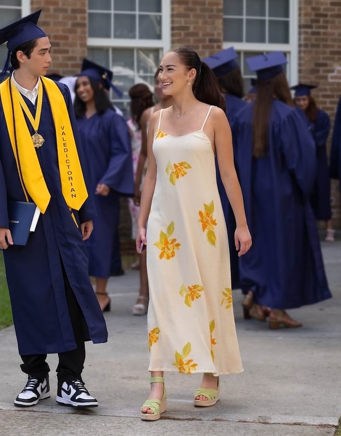 A young woman in a long white dress with yellow flowers, inspired by The Summer I Turned Pretty outfits, smiles while walking outside at a graduation ceremony. Nearby, a graduate in a blue cap and gown with a yellow Valedictorian sash holds a diploma.