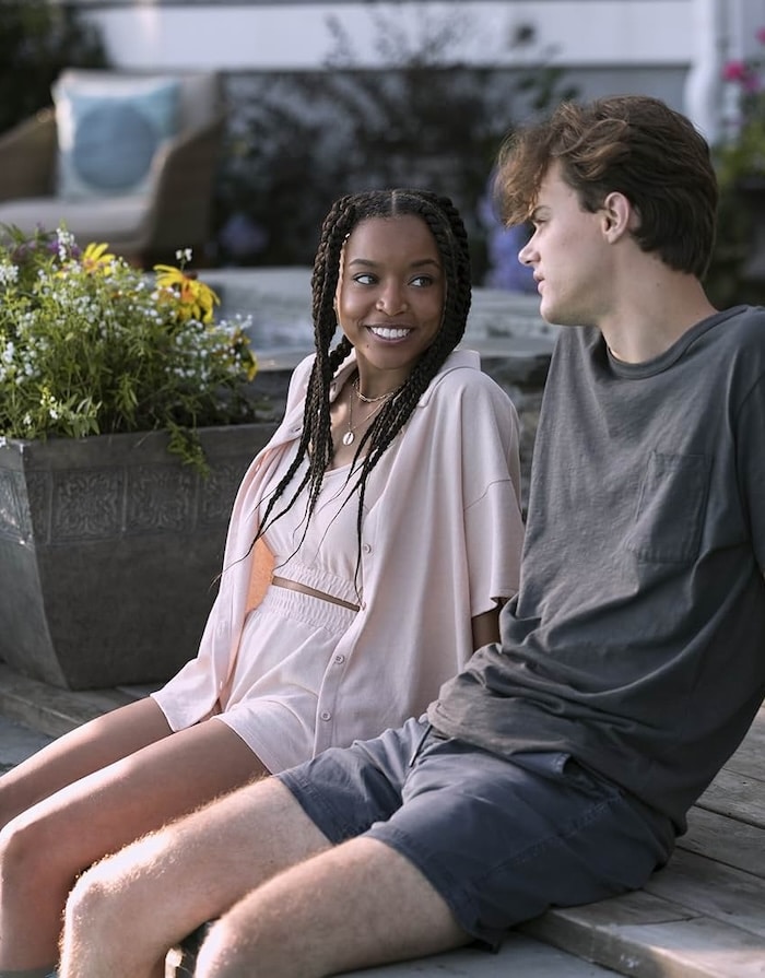 Two young people sit on outdoor steps, smiling at each other. The woman rocks braided hair and a light pink summer fashion look, while the man wears a dark t-shirt and shorts. A planter with flowers is visible behind them.