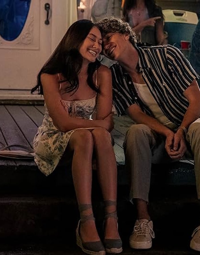 A young woman and man sit close together on porch steps at night, smiling and leaning their heads toward each other in The Summer I Turned Pretty-inspired outfits—a floral dress with wedges for her, a striped shirt and sneakers for him.