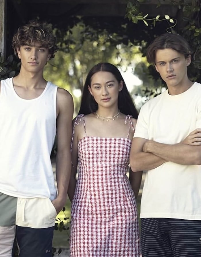 Three young adults, dressed in trendy summer outfits, stand side by side outdoors—channeling the carefree vibe of The Summer I Turned Pretty. One wears a white tank top and shorts, another a red plaid dress, and the third sports a white t-shirt with arms crossed.