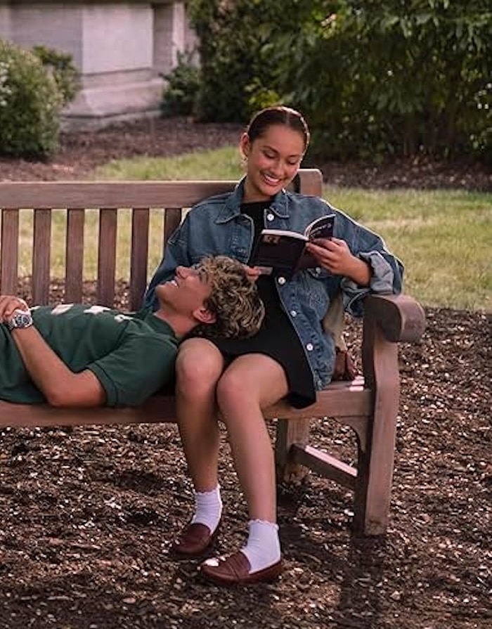 Two people sit on a wooden bench outdoors; one, dressed in stylish summer outfits, rests their head on the other’s lap while the seated person smiles and reads a book. Trees, grass, and a building appear in the background.
