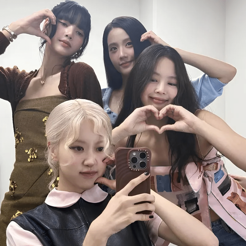 Four young women pose together for a selfie in a well-lit room, making heart shapes with their hands and smiling at the camera, capturing the excitement of BLACKPINK tour prep. One holds a phone to take the photo.
