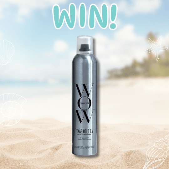 A silver can of Color Wow Texas Hold Em Hairspray stands upright on sandy beach with a blurred ocean and palm trees in the background. The word WIN! appears above the can in large, playful letters.