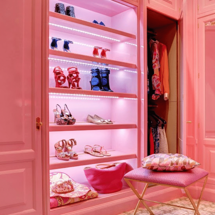 A brightly lit, pink-themed closet displays colorful high-heeled shoes on shelves, with handbags below. A pink stool with patterned pillows sits in front, and hanging clothes are visible in the open wardrobe.