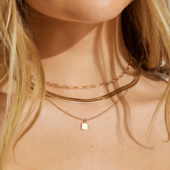 A close-up of a womans neck wearing three layered gold necklaces, one with a small square pendant. Her light skin and blonde hair drape over her shoulders, adding a touch of The Summer I Turned Pretty style to her outfits.