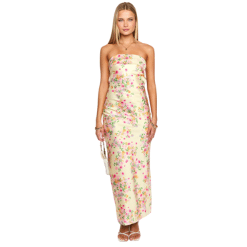 A woman stands wearing a strapless, floor-length cream dress with a pink and green floral pattern, holding a small white handbag and wearing white sandals—an inspired outfit perfect for fans of The Summer I Turned Pretty.